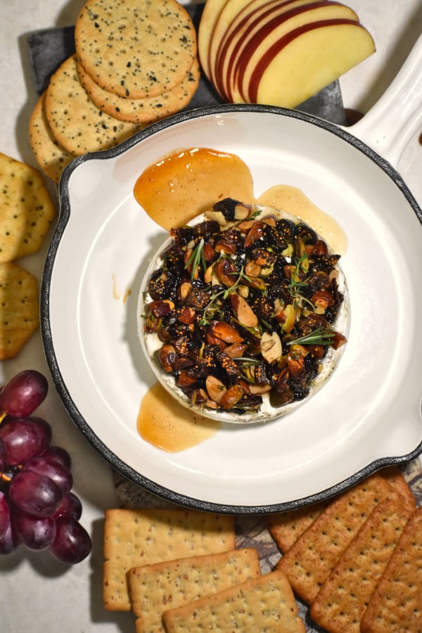 baked brie with nuts and honey in a small cast iron pan surrounded by crackers and fruit